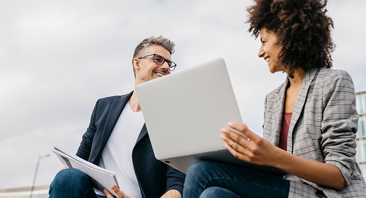 A man and woman are sitting on a bench looking at a laptop.